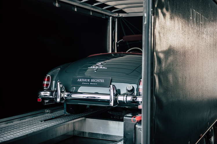 Ein klassisches graues Auto steht in einem Transportanhänger. Die Heckansicht zeigt glänzende Chromdetails und das Logo. Ein Schild mit der Beschriftung Arthur Bechtel Classic Motors ist gut sichtbar. Der Hintergrund ist dunkel, wodurch das Auto hervorsticht.