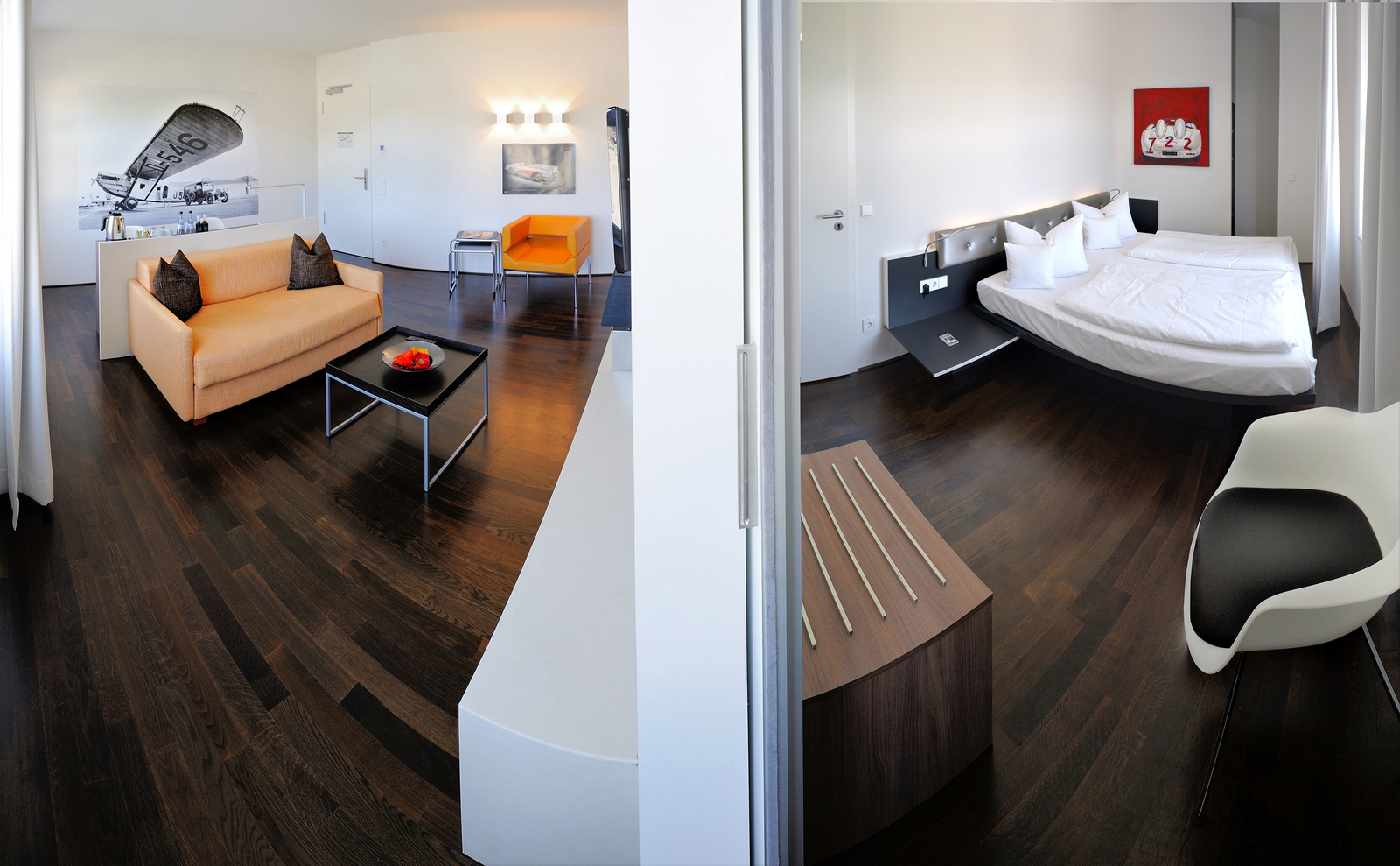 Das Bild zeigt ein modernes Zimmer mit einem Wohnbereich auf der linken Seite. Dort befindet sich ein orangefarbenes Sofa, ein schwarzer Couchtisch mit einer Vase und Stühlen. Auf der rechten Seite ist der Schlafbereich mit einem Doppelbett und einem abstrakten Kunstwerk an der Wand. Der Boden ist aus dunklem Holz.