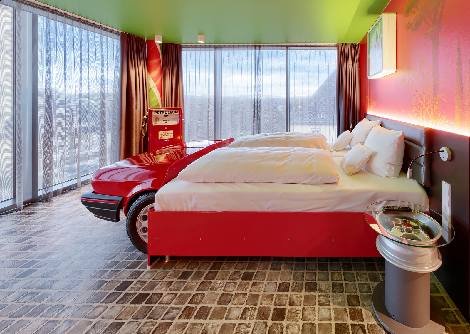 Ein modernes Hotelzimmer mit buntem Design. Ein Doppelbett mit weißen Kissen steht neben einem roten Auto als Kopfteil. An der Wand befindet sich eine große Fensterfront mit Vorhängen, die viel Licht hereinlässt, und ein stilisierter Zapfsäulen-Automat. Der Boden hat einen hübschen Steinbelag.