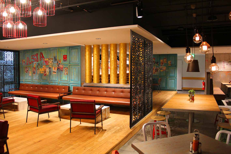 Ein modernes Restaurantinterieur mit Holzfußboden und verschiedenen Sitzbereichen. An der Wand hängen bunte Plakate, während goldene Säulen und stilvolle Lampen das Ambiente beleben. Gemütliche Sofas und Tische laden zum Verweilen ein.