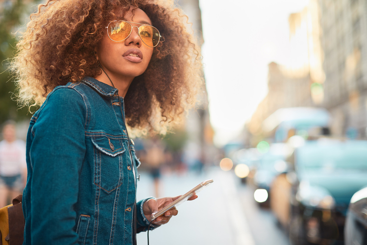 Eine junge Frau mit lockigem Haar trägt eine Jeanjacke und Sonnenbrille. Sie steht auf einer belebten Straße und schaut nachdenklich in die Ferne, während sie ein Smartphone in der Hand hält. Im Hintergrund sind verschwommene Autos und Passanten zu sehen.