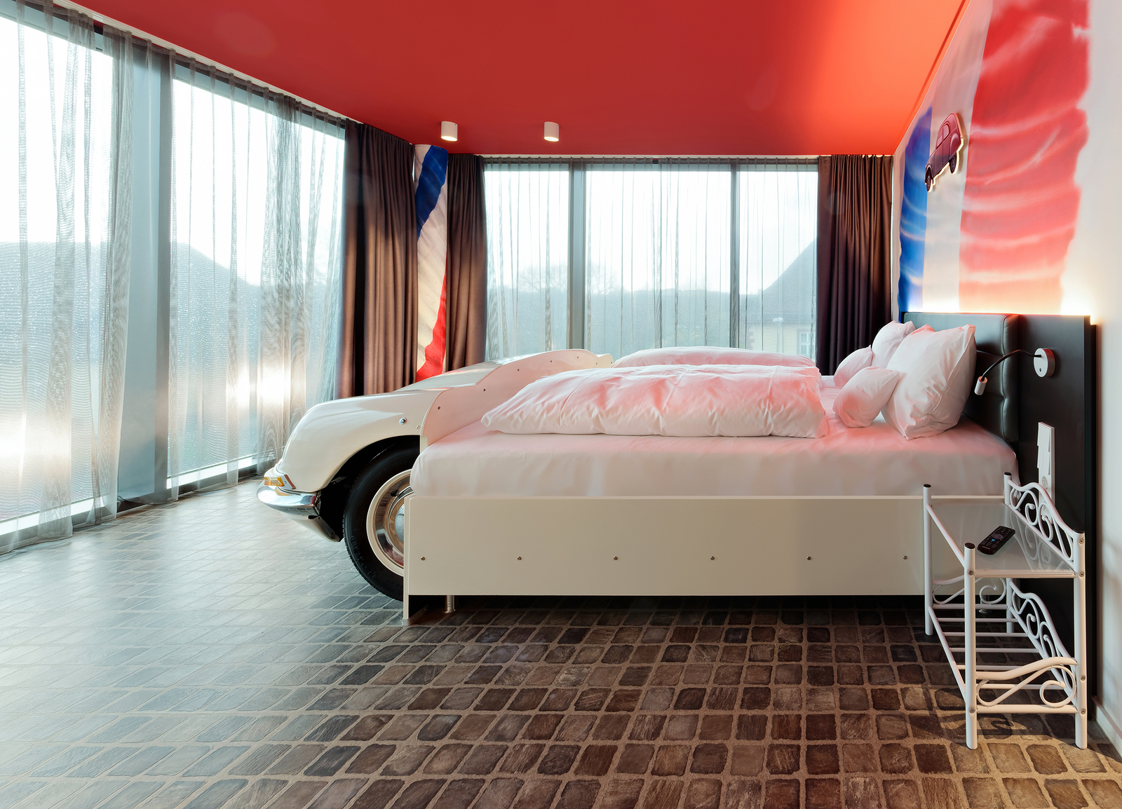 Das Bild zeigt ein modernes Schlafzimmer mit einem Bett, das an ein Auto erinnert. Neben dem Bett steht eine detaillierte Autobodeneinheit. Große Fenster lassen viel Licht herein und der Raum hat eine leuchtend rote Decke. Der Fußboden ist in Steinoptik gestaltet.