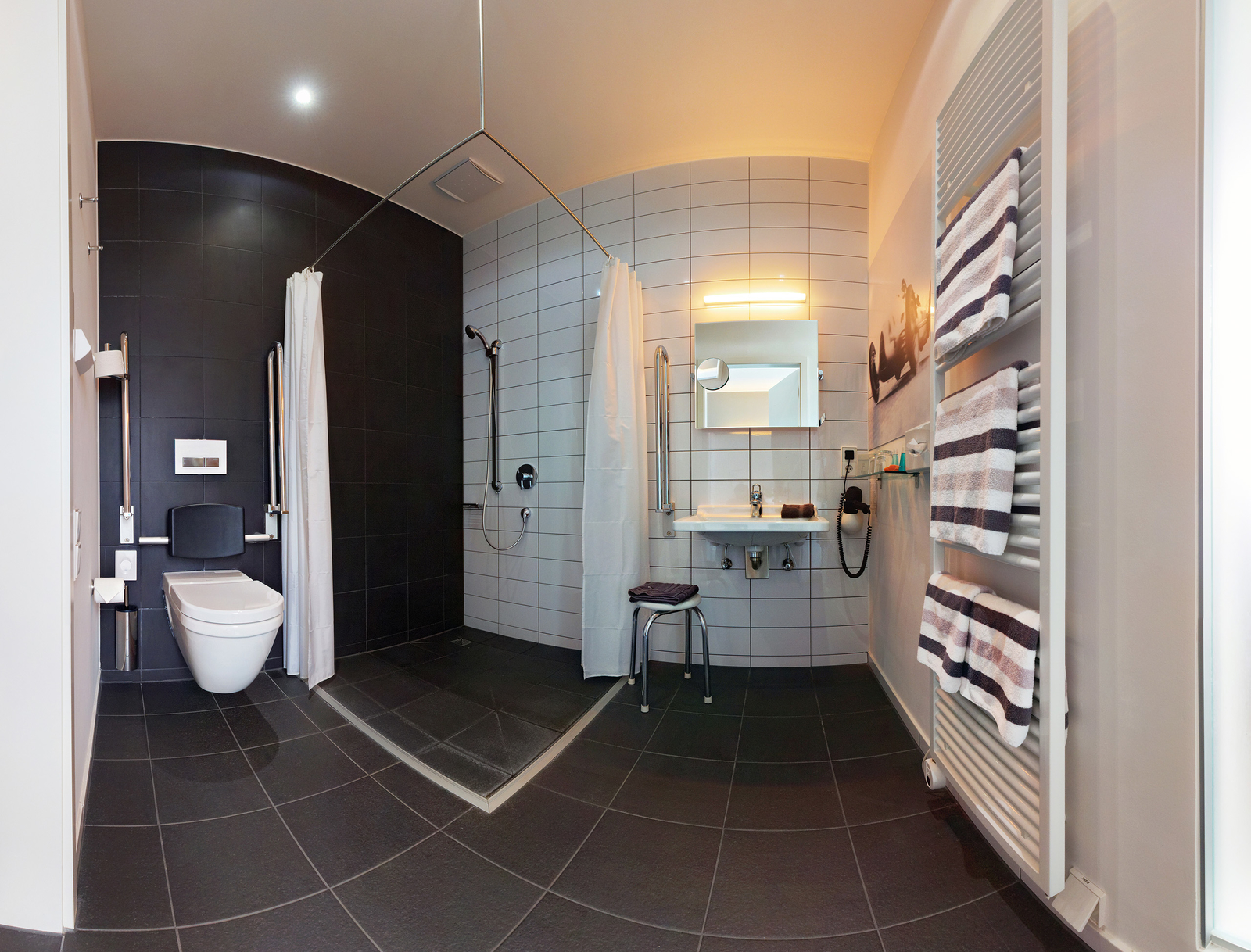 Das Bild zeigt ein modernes, barrierefreies Badezimmer mit einer Dusche und einer Toilette. An der Wand sind Handtücher aufgehängt, und ein Waschbecken steht auf einem kleinen Tisch. Die Einrichtung ist durch klare Linien und eine helle Farbpalette gekennzeichnet, was einen sauberen und funktionalen Eindruck vermittelt.