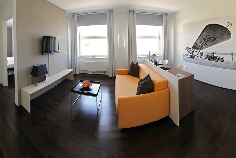Das Bild zeigt ein modernes Zimmer mit einem orangenen Sofa, einem Tisch mit Obstschale und einem Fernseher an der Wand. Große Fenster sorgen für viel Tageslicht. An der Wand ist ein historisches Flugzeugmotiv abgebildet, während der Boden aus dunklem Holz besteht.