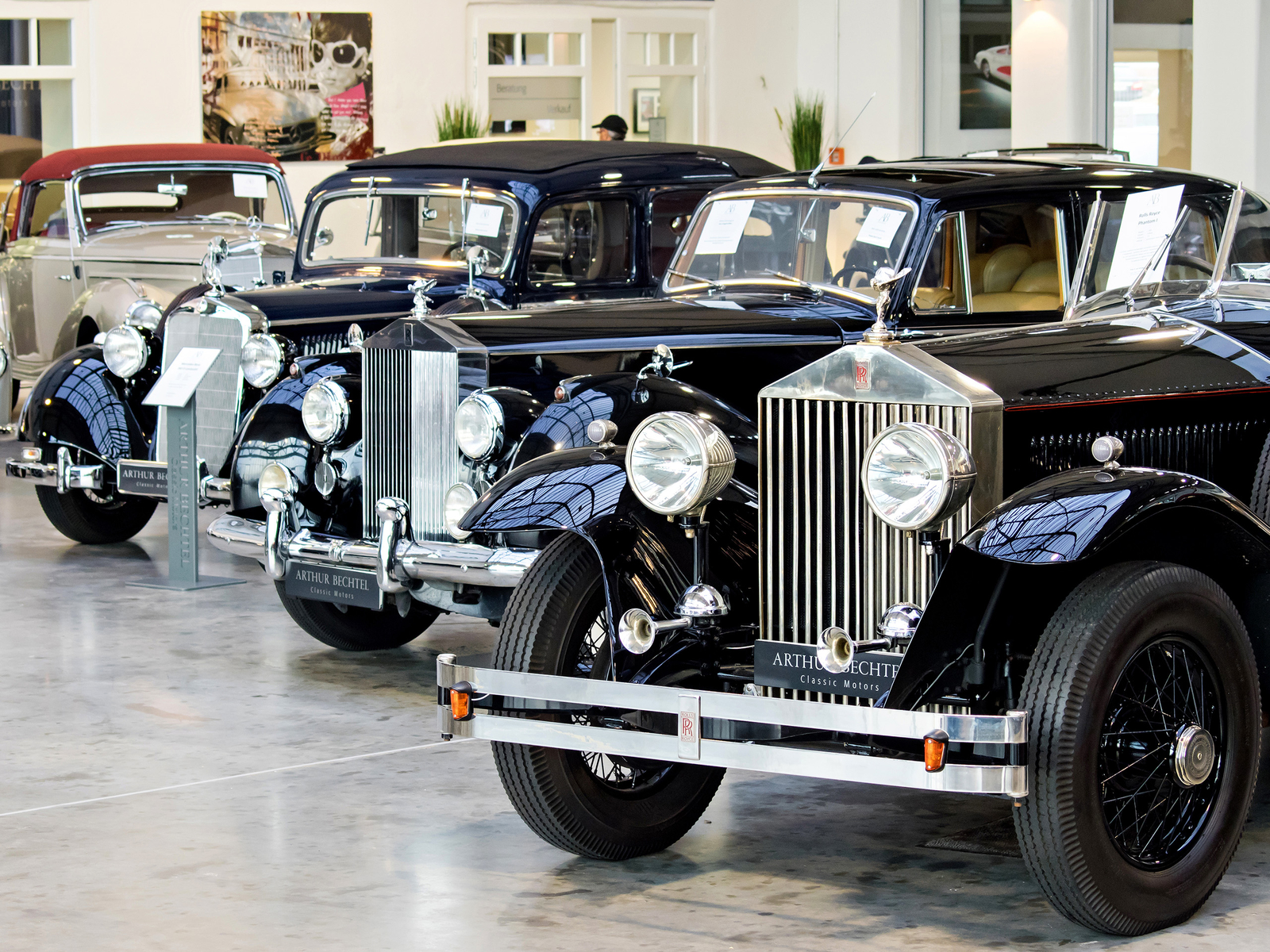 Die Abbildung zeigt mehrere klassische schwarze Rolls-Royce-Autos in einer Ausstellungshalle. Die Fahrzeuge sind in einer Reihe angeordnet, wobei ihre eleganten Designs und Chromdetails gut zur Geltung kommen. Auf einzelnen Autos sind Beschilderungen angebracht, die Informationen zu den Modellen bieten.
