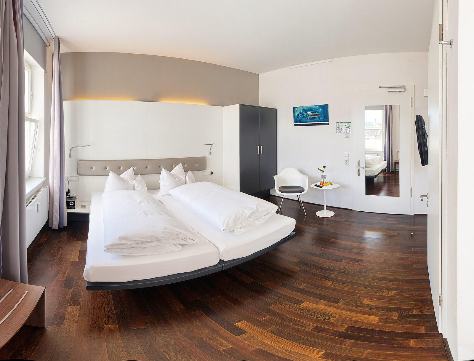 Das Bild zeigt ein modernes Hotelzimmer mit einem Doppelbett und zwei separaten Matratzen. An der Wand sind Bilder und zwei Fenster mit Vorhängen. Ein kleiner Tisch mit einer Blume und einer Snackplatte befindet sich im Raum. Der Boden ist aus Holz.