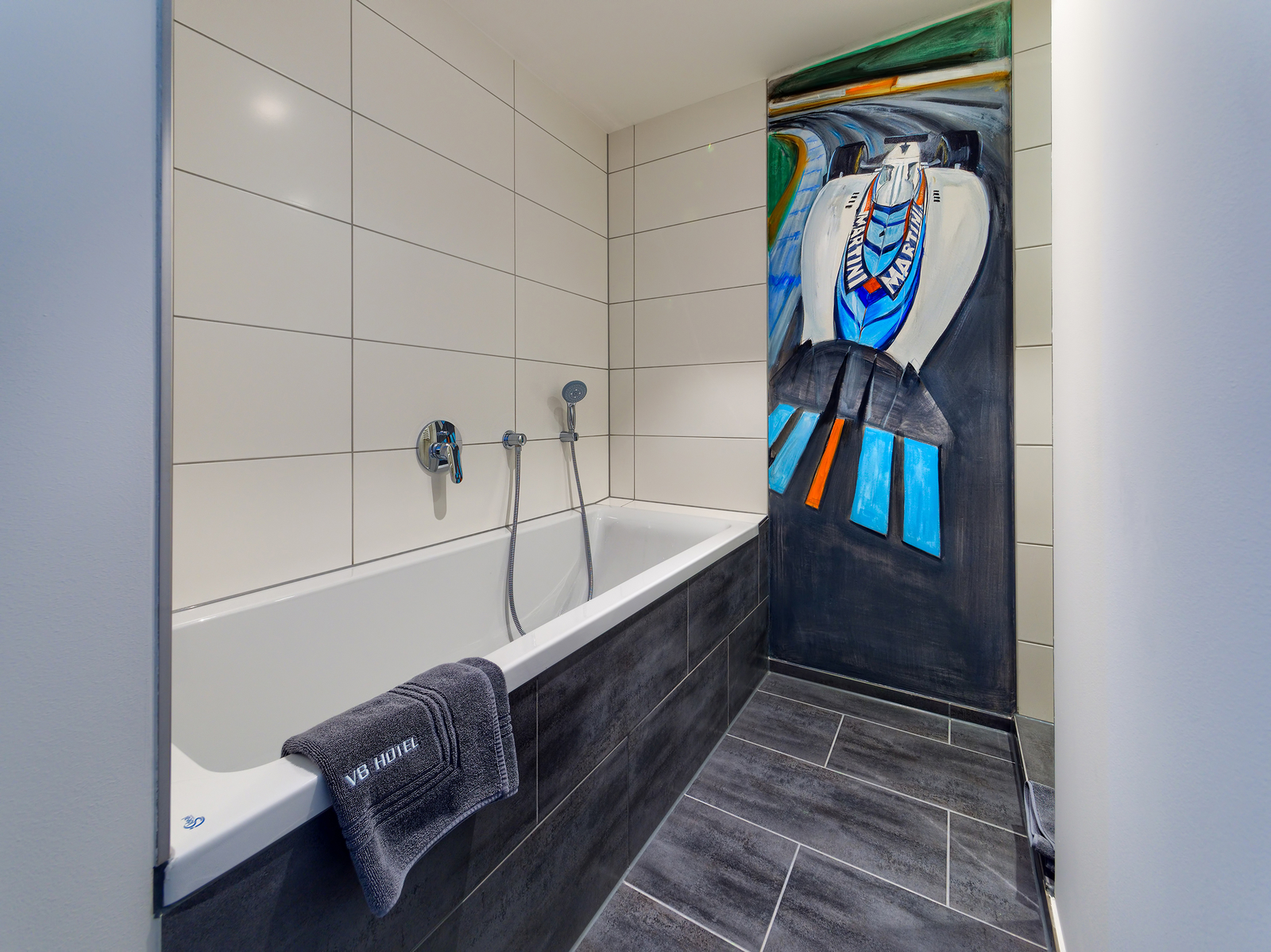 Das Bild zeigt ein modernes Badezimmer mit einer Badewanne und einer Handbrause. An der Wand befindet sich ein farbenfrohes, künstlerisches Wandgemälde eines Rennwagens. Der Boden ist mit großen, dunklen Fliesen ausgelegt und ein Handtuch mit dem Aufdruck „V8 HOTEL“ liegt auf der Badewanne.