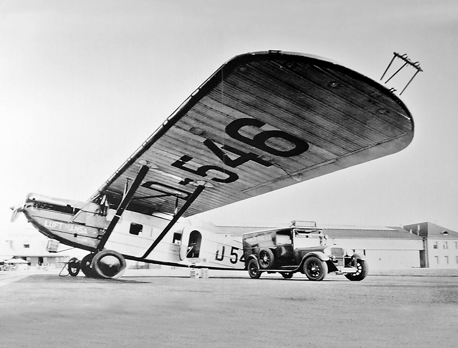 Das Bild zeigt ein historisches Flugzeug mit dem Schriftzug "LUFTHANSA" und der Nummer 546, das auf einem Flugplatz steht. Ein altes Auto ist neben dem Flugzeug positioniert. Die Aufnahme ist in Schwarz-Weiß gehalten und vermittelt einen Eindruck von der Luftfahrt vergangener Zeiten.