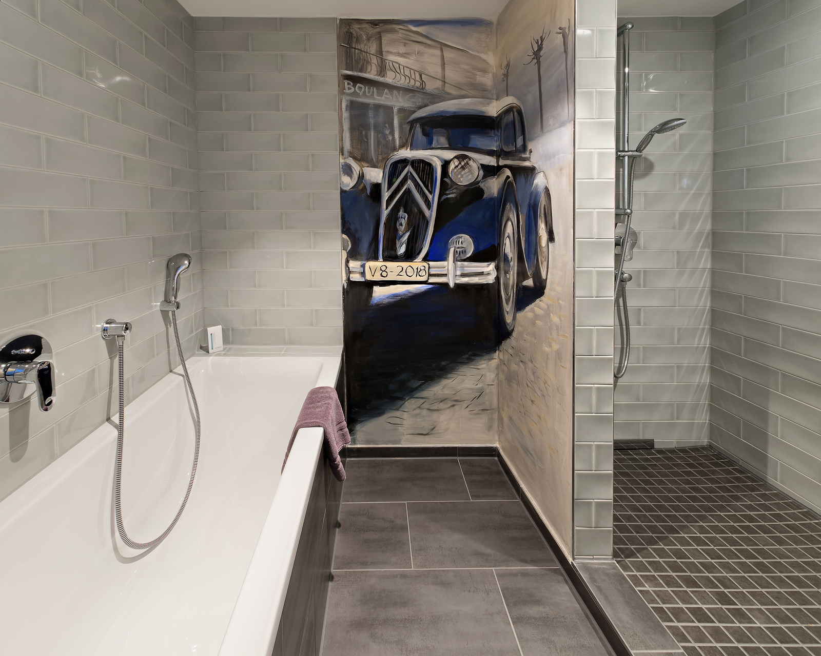 Ein modernes Badezimmer mit einer Badewanne und einer Dusche. An einer Wand ist ein großes Wandbild eines alten blauen Autos, das auf einer Straße steht. Die Wände sind mit grauen Fliesen gefliest, und ein Handtuch liegt auf der Badewanne.