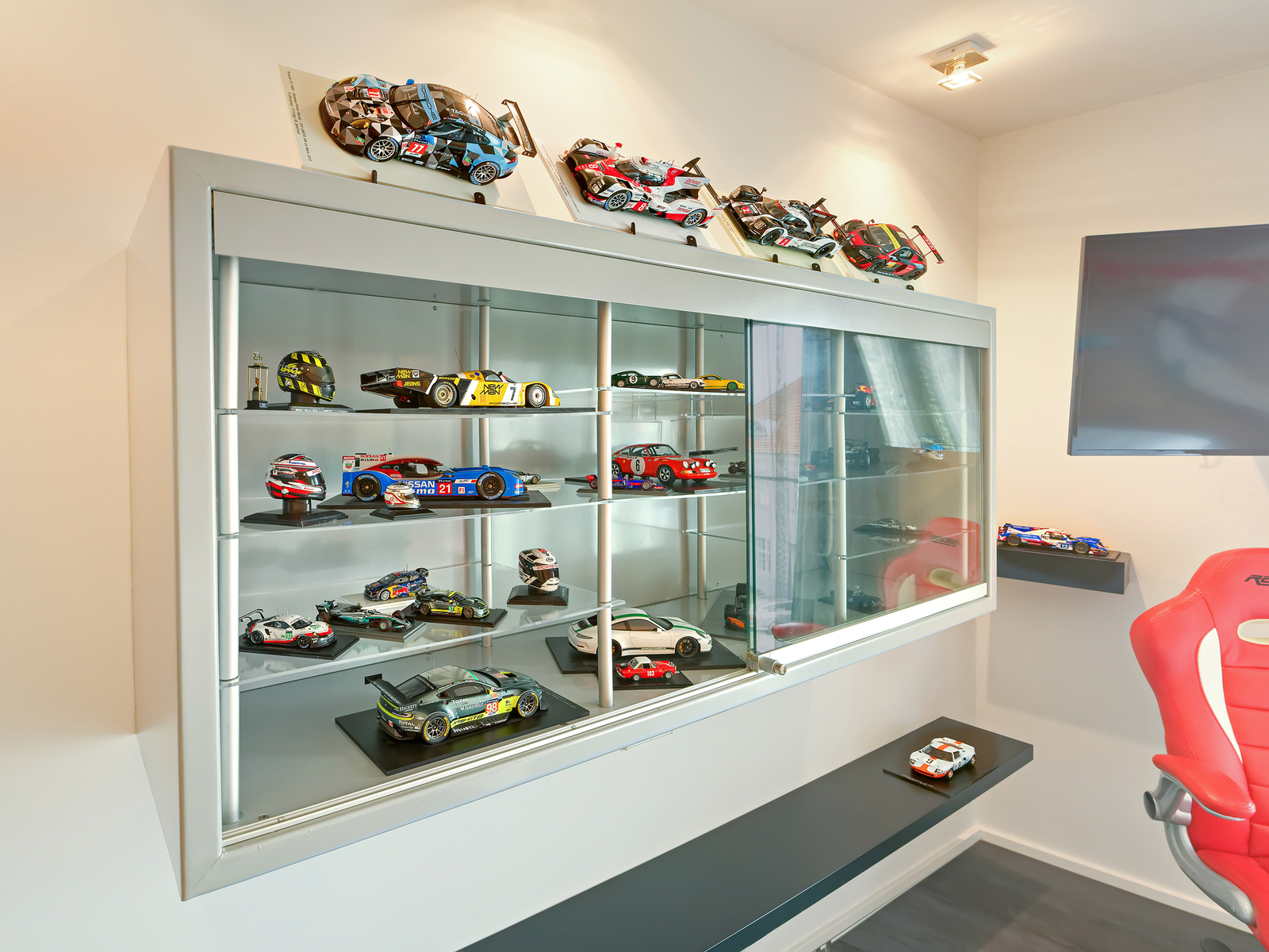 Ein Glasschrank mit mehreren Modellautos, darunter Rennwagen, ist abgebildet. Oben sind einige weitere Modelle und Helme platziert. Der Schrank hat mehrere Regale, die somit eine Vielzahl an Autos aus unterschiedlichen Perspektiven zeigen. Das gesamte Arrangement vermittelt eine Rennsport-Atmosphäre.