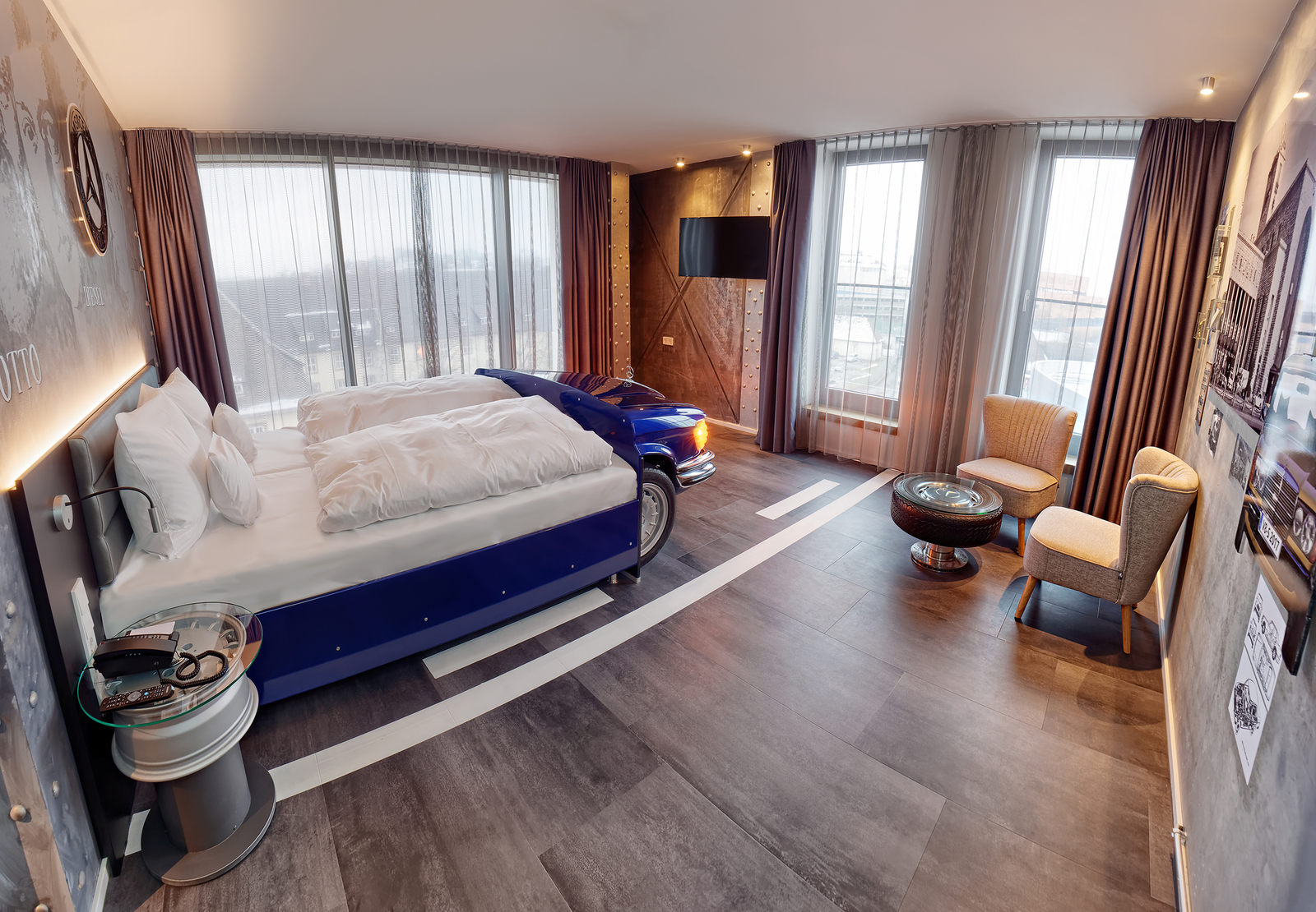 Das Bild zeigt ein modernes Hotelzimmer mit einem großen Doppelbett, ausgestattet mit weißen Bettdecken. An einer Wand steht ein blauer, automobilähnlicher Möbelstück, während weitere Sitzgelegenheiten und ein Tisch im Raum platziert sind. Große Fenster sorgen für viel Tageslicht.
