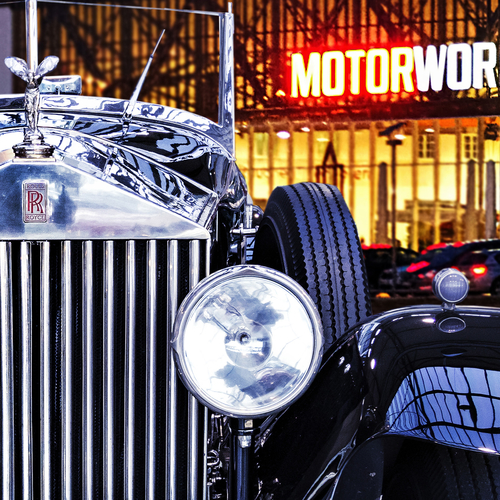 Ein klassischer Rolls-Royce mit glänzender Motorhaube und elegantem Kühlergrill ist abgebildet. Das Fahrzeug hat eine runde Scheinwerfer und eine dekorative Figur auf der Haube. Im Hintergrund sind die beleuchteten Fenster eines modernen Gebäudes zu sehen, auf dem "MOTORWORLD" steht.