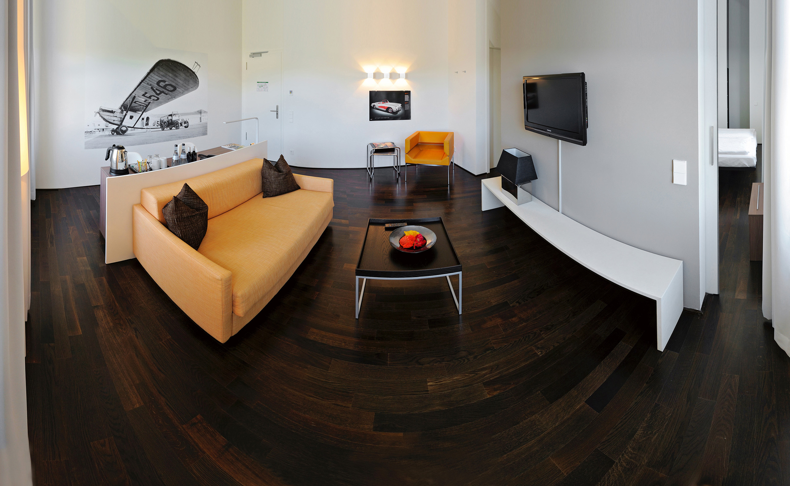 In einem modernen Zimmer sind ein oranges Sofa mit Kissen, ein schwarzer Tisch mit einer Obstschale, ein Fernseher an der Wand und ein helles Stuhl vorhanden. Der Boden ist aus dunklem Holz, und an der Wand hängen Fotografien. Die Raumgestaltung ist minimalistisch und stilvoll.
