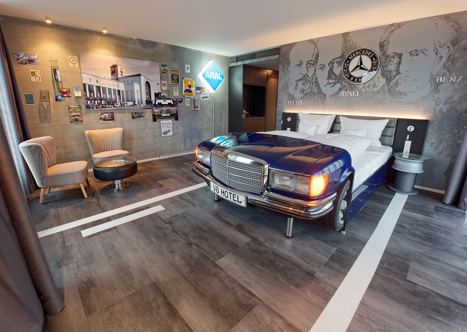 Ein Schlafzimmer im V8 Hotel, das aussieht wie ein Auto. Ein blauer Mercedes als Bett, mit einem Tisch aus einem Autoreifen und stilvollen Stühlen. Die Wände sind mit historischen Fotos und Bildern von Automobilpionieren dekoriert.