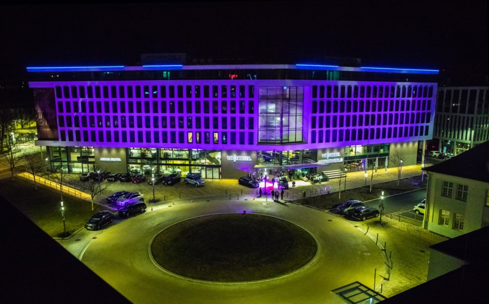 Eine moderne, mehrstöckige Gebäudeansicht bei Nacht, beleuchtet in lila und blau. Vor dem Gebäude parken Autos, und es sind einige Personen vor dem Eingang versammelt. Der Rundweg vor dem Gebäude ist gut beleuchtet.