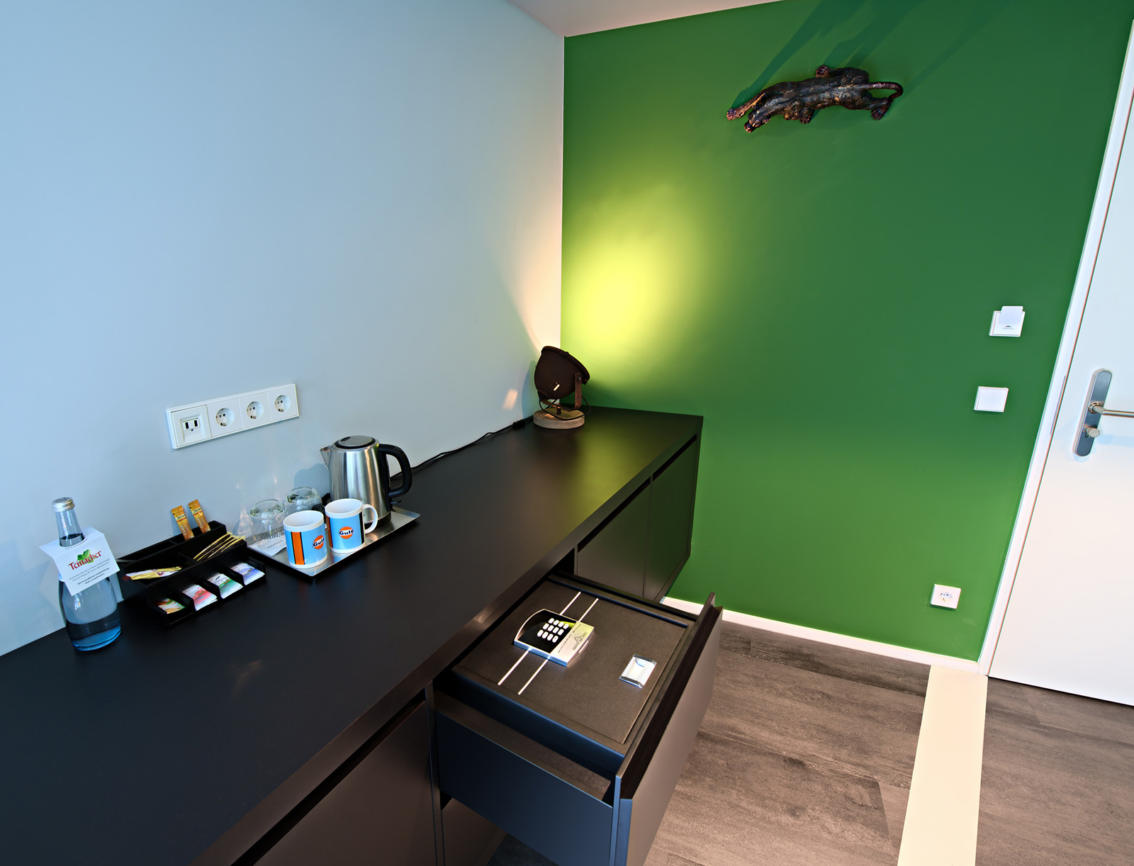 Das Bild zeigt eine moderne Ecke eines Raumes mit einem schwarzen Schreibtisch. Auf dem Tisch stehen eine Kaffeekanne, zwei Tassen, ein Wasserglas und ein Tablett mit Teebeuteln. Eine grüne Wand und eine aufgeräumte Schublade mit einem Zahlenschloss sind sichtbar.