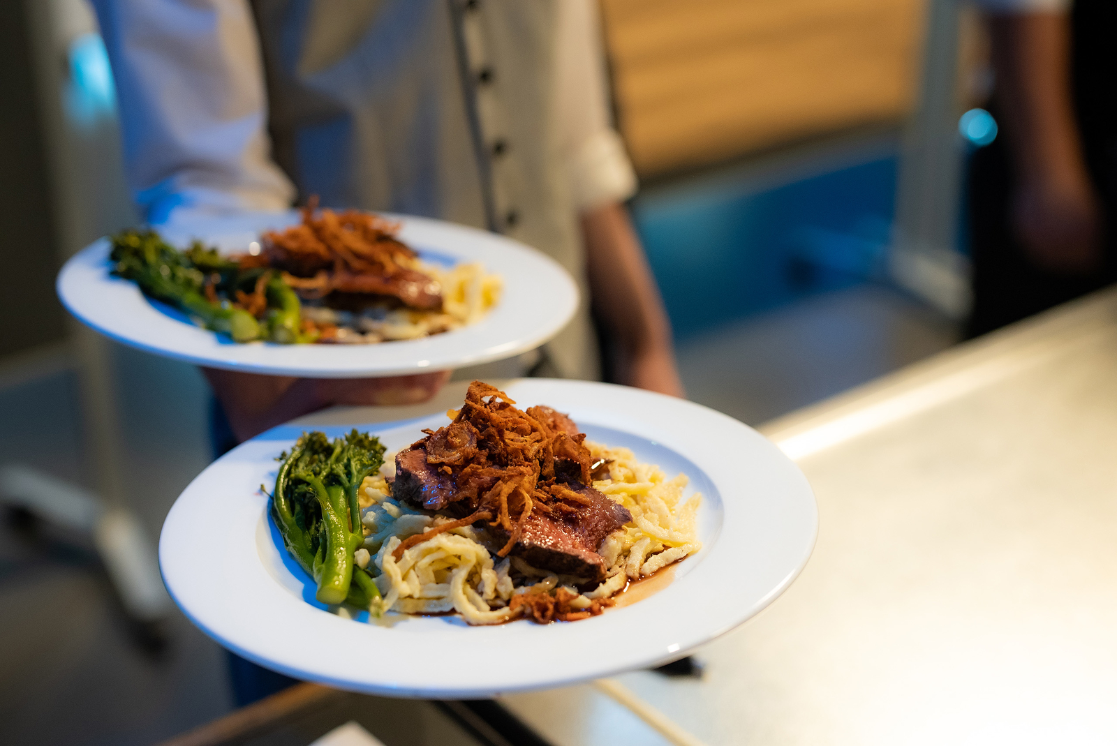 Zwei weiße Teller mit einer Portion Fleisch, frittierten Zwiebeln, Spätzle und Brokkoli werden von einer Hand serviert. Die Speisen sind ansprechend angerichtet und scheinen frisch zubereitet zu sein, während ein unscharfer Hintergrund ein Restaurantambiente vermittelt.