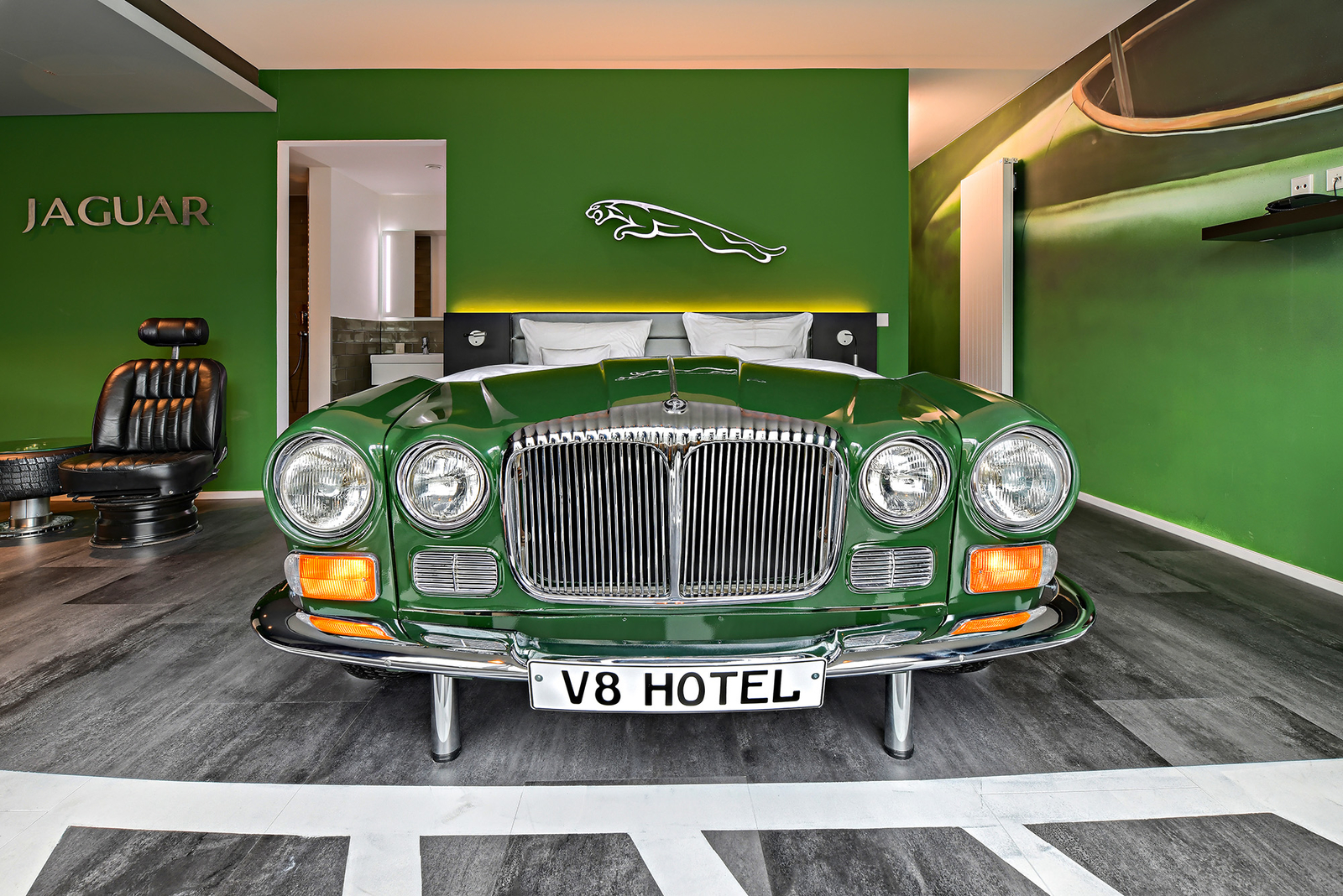 Grünes Autobett neben einem schwarzem Sessel aus einem Autositz im Jaguar-Themenzimmer im V8 Hotel an einer grünen Wand mit silbernem Jaguar-Logo