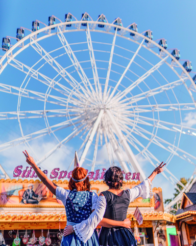 Zwei Personen in traditioneller Tracht stehen Arm in Arm vor einem großen Riesenrad. Sie zeigen ein Peace-Zeichen mit einer Hand. Der Himmel ist blau und die Stimmung wirkt fröhlich und ausgelassen, was auf einen Festplatz hinweist.