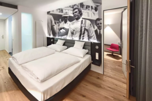 Doppelzimmer mit Holzboden und großflächigem Schwarz-Weiß-Fotomotiv eines Rennfahrers im Gulf-Rennoverall hinter dem Bett, rechts Blick in ein Nebenzimmer mit rotem Stuhl.