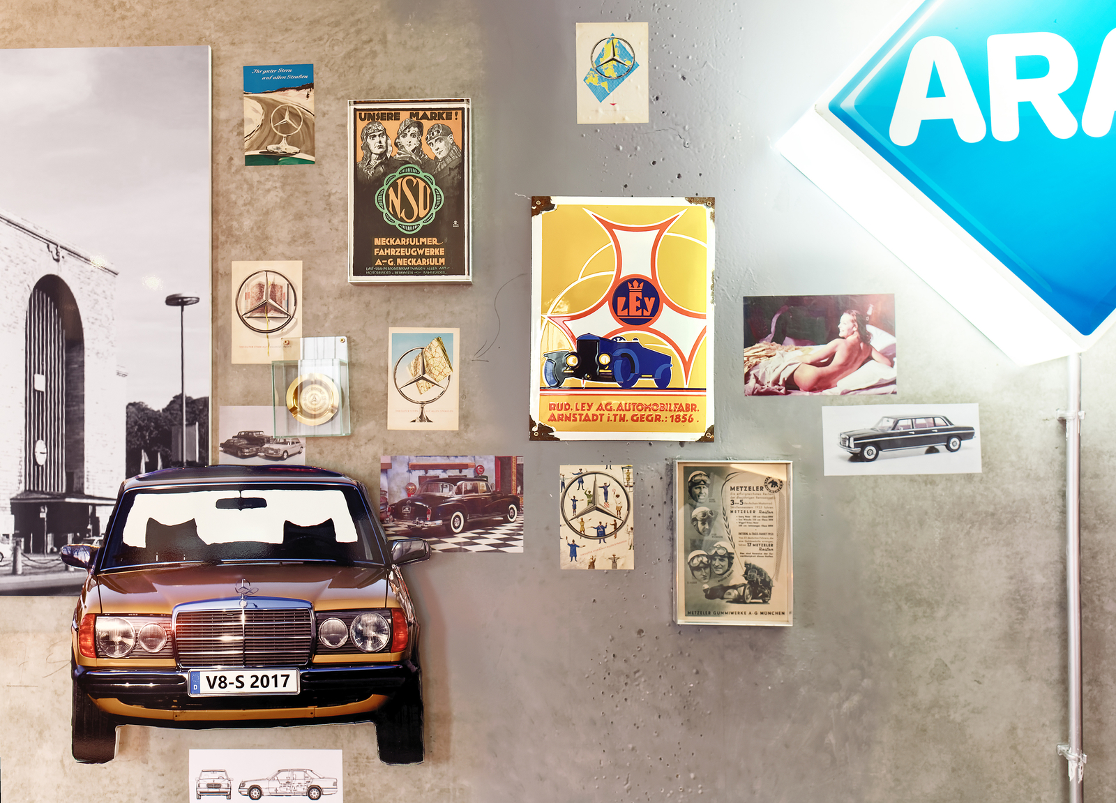 Das Bild zeigt ein schwarzes Auto, das zentral an einer Wand hängt, umgeben von verschiedenen historischen Autoplakaten und Fotos. Aus einer Neonreklame leuchtet das Wort "ARAL". Die Wände sind in einem schlichten, grauen Farbton gehalten.