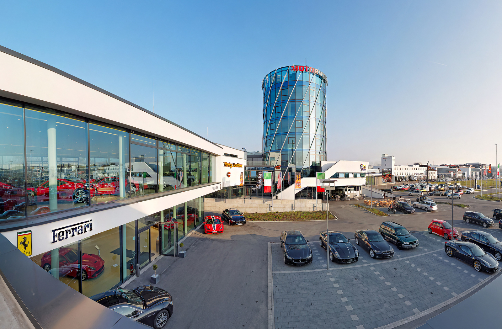 Das Bild zeigt ein modernes Autohaus mit einer großen Glasfassade, in der mehrere Ferrari-Modelle ausgestellt sind. Im Hintergrund steht ein beeindruckender, gläserner Turm mit der Aufschrift Motorworld. Vor dem Gebäude sind verschiedene Autos auf einem Parkplatz zu sehen.