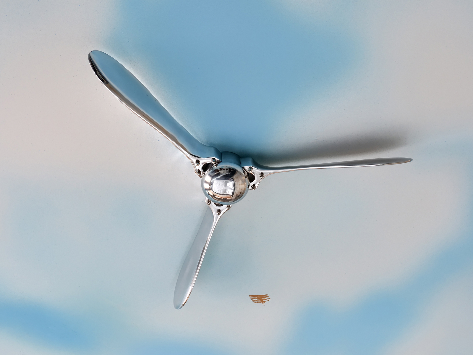 Bildbeschreibung: Ein Deckenventilator mit drei metallischen Blättern und einem runden, glänzenden Motor in der Mitte, vor einem hellblauen, leicht strukturierte Hintergrund, der an einen Himmel erinnert. Die Blätter sind elegant und modern gestaltet.
