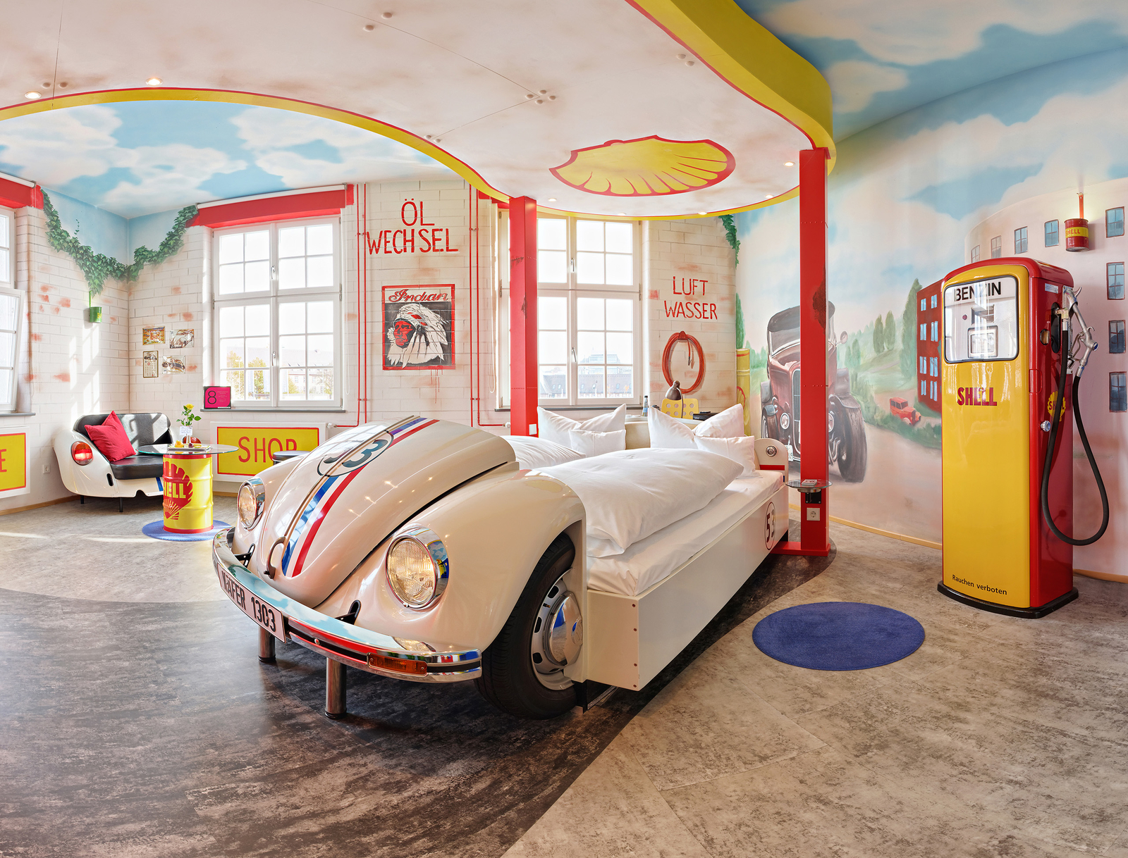 Ein stilvolles Zimmer im Retro-Design mit einem Bett, das wie ein klassisches Auto aussieht, und einer nostalgischen Tanksäule. Wände sind mit Fahrzeugmotiven und bunten Akzenten dekoriert. Große Fenster sorgen für viel Licht und Frische. Ideal für Auto-Enthusiasten und Liebhaber des Vintage-Stils.
