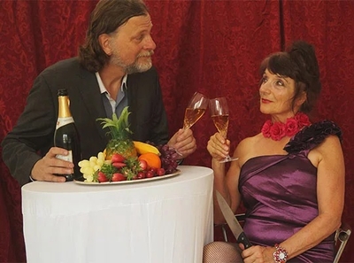 Ein Mann und eine Frau stoßen mit Gläsern an. Der Mann trägt einen Anzug und hält eine Flasche Sekt, während die Frau in einem eleganten lila Kleid mit einer Blume am Hals sitzt. Auf dem Tisch liegt eine Obstplatte. Sie wirken fröhlich und feiern einen besonderen Moment.