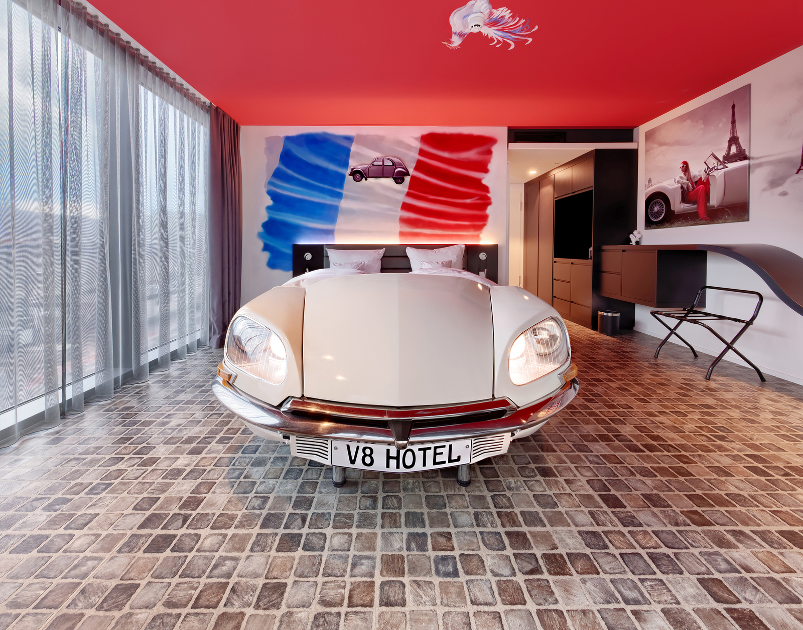 Das Bild zeigt ein stilvolles Hotelzimmer mit einem einzigartigen Design. Im Mittelpunkt steht ein Bett, das wie ein klassisches Auto gestaltet ist. Der Hintergrund ist mit französischen Flaggenfarben und einer Eiffel-Turm-Dekoration verziert. Große Fenster bieten viel Tageslicht.