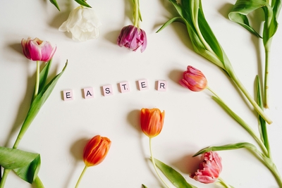 Auf einer weißen Fläche liegen verschiedene Tulpen in unterschiedlichen Farben, umgeben von grünen Blättern. In der Mitte steht das Wort "EASTER" aus pinken Buchstabenschnitten.