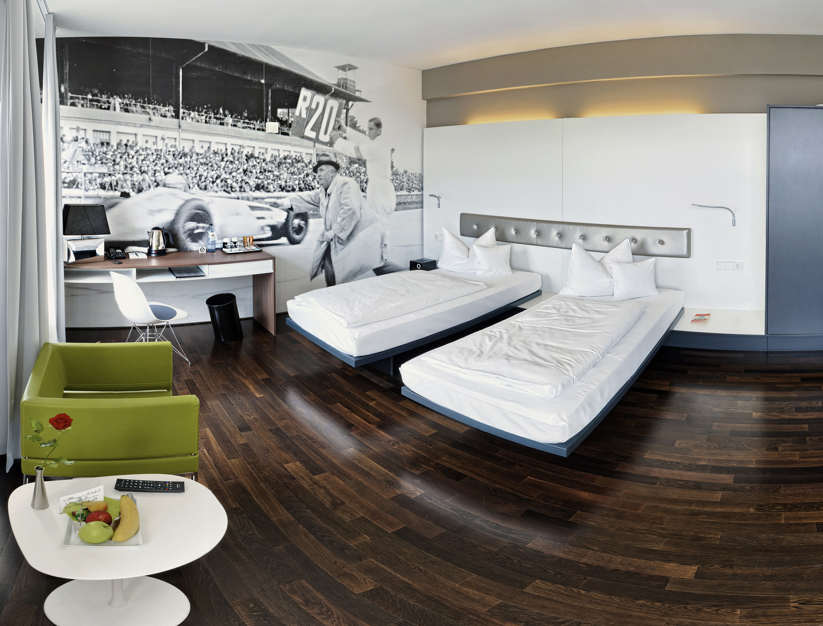 Ein modernes Hotelzimmer mit zwei Einzelbetten, elegant und minimalistisch eingerichtet. In der Ecke steht ein grüner Sessel. Auf dem Tisch befinden sich eine Schale mit Obst, eine Fernbedienung und eine Blume in einer Vase. An der Wand ist ein großes historisches Foto einer Rennszene zu sehen.