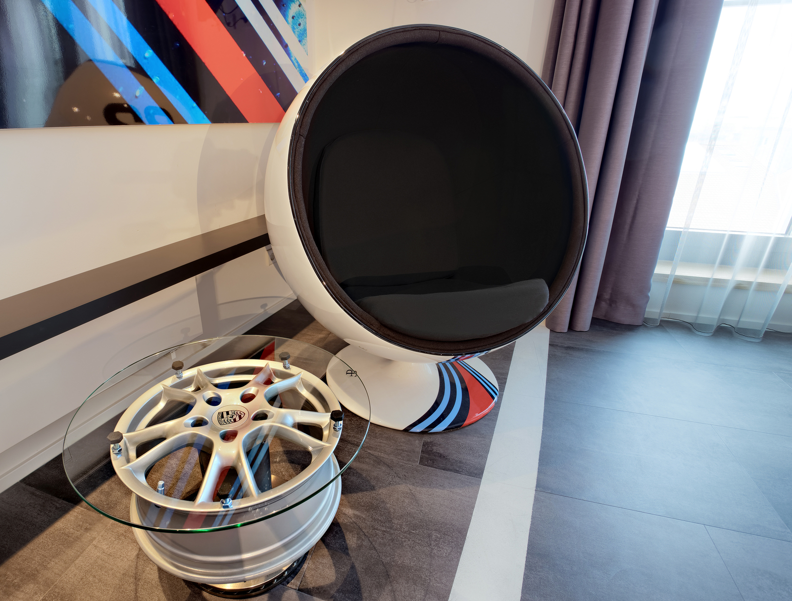 Ein moderner, runder Sessel mit einem schwarzen Innenbereich steht neben einem Glas- und Aluminium-Tisch, der aus einer Autoreifenfelge besteht. Die Wand dahinter ist mit einem bunten, abstrakten Kunstwerk dekoriert. Vor dem Fenster hängen lichte Vorhänge.