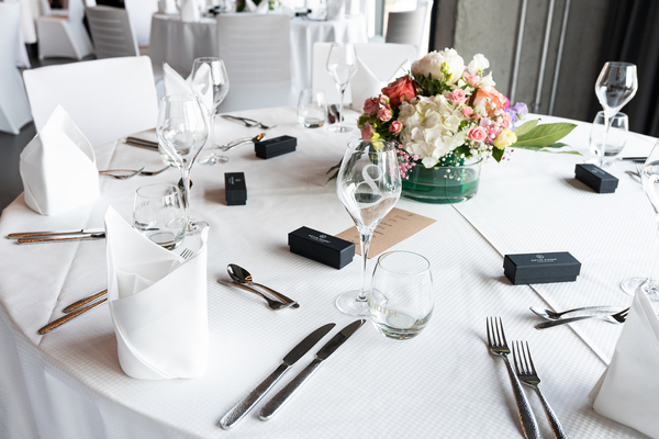 Ein elegant gedeckter Esstisch mit weißen Servietten, Gläsern und versilbertem Besteck. In der Mitte befindet sich ein blumiges Arrangement mit rosa und weißen Blumen in einer Glasvase. Schwarze Boxen liegen um den Tisch verteilt.