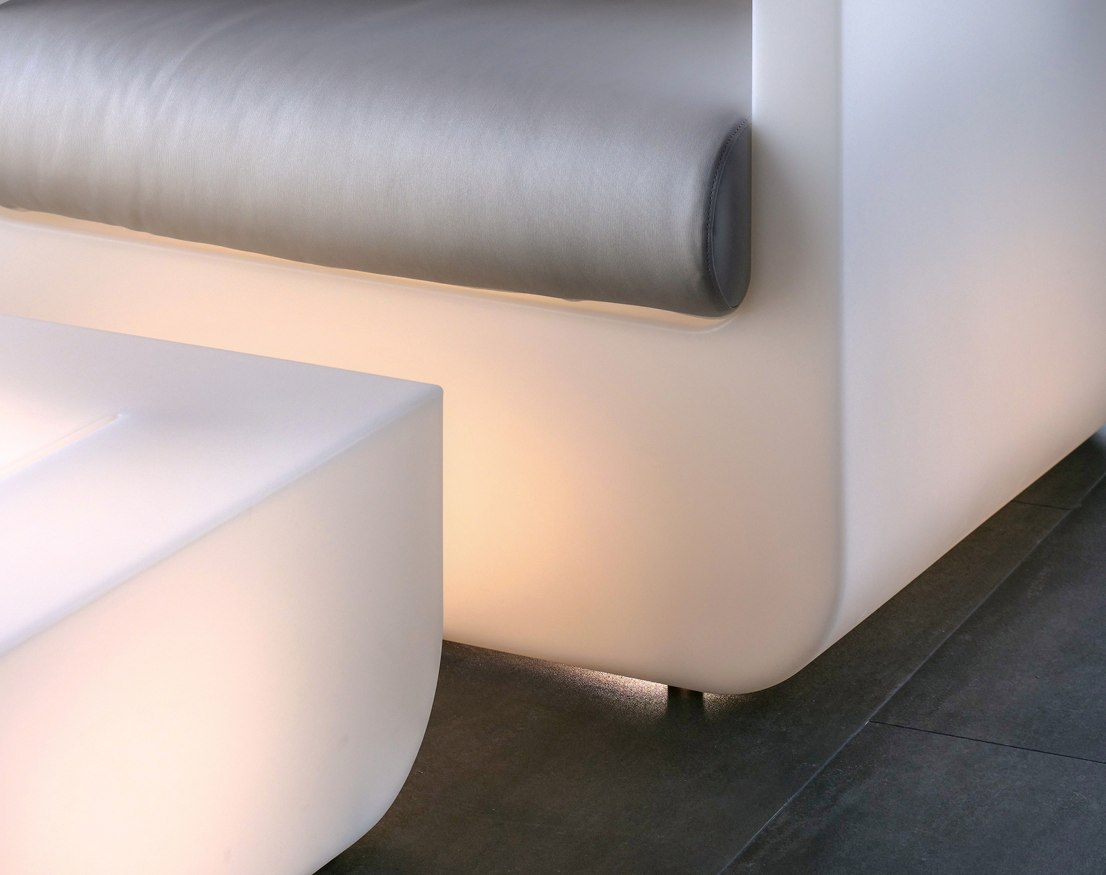 Eine moderne, minimalistische Sofakonstruktion mit einem grauen Kissen und sanfter, indirekter Beleuchtung an den Kanten. Die klaren, runden Formen und die hellen Farben schaffen eine elegante Atmosphäre in einem Wohnraum.
