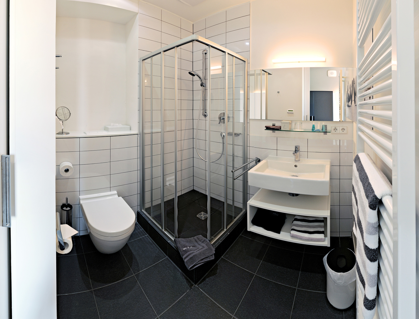 Das Bild zeigt ein modernes Badezimmer mit einer Dusche, einem Waschbecken und einer Toilette. Die Wände sind hell gefliest, und es gibt einen Handtuchhalter sowie einen Abfalleimer. Die Dusche ist mit einer Glasabtrennung versehen und die Ausstattung wirkt sauber und funktional.