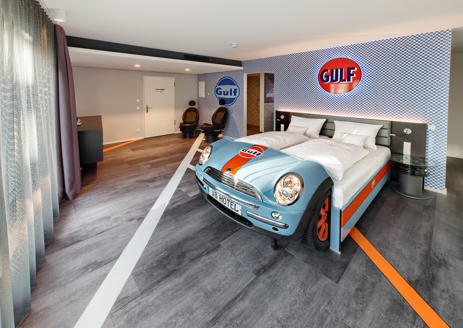 Gulf-Themenzimmer im V8 Hotel mit einem hellblauen Autobett an einer karierten Wand neben zwei Autositzsesseln