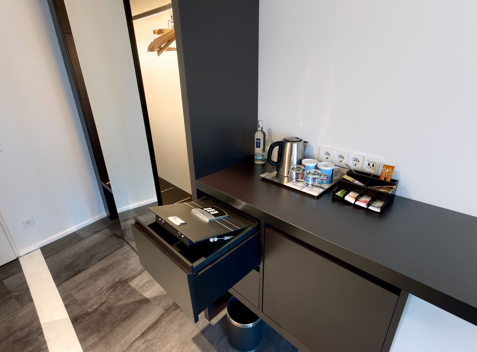In einem modernen Hotelzimmer sieht man einen schwarzen Schreibtisch mit einem offenen Schublade, die einen Safe zeigt. Auf dem Tisch stehen eine Teekanne, Tassen, ein Wasserglas und verschiedene Tee- und Kaffeepackungen. Im Hintergrund ist ein Kleiderschrank sichtbar.