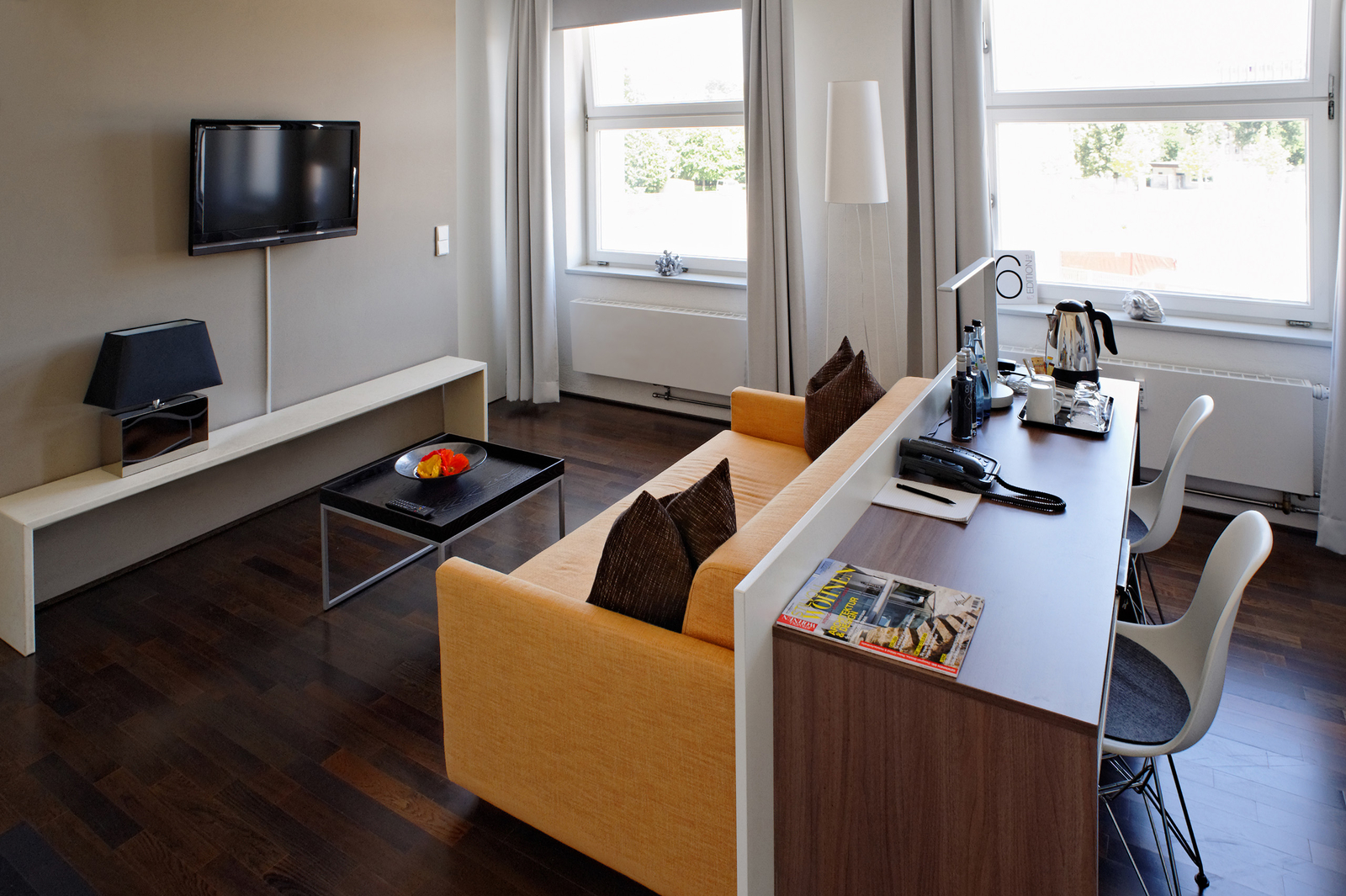 Ein modernes Wohnzimmer mit einem orangen Sofa, einem Tisch mit einer Schale frischem Obst und einem Fernseher an der Wand. Auf einem Schreibtisch stehen Getränke, Gläser und Schreibutensilien. Große Fenster lassen Licht herein und bieten einen Blick ins Freie.