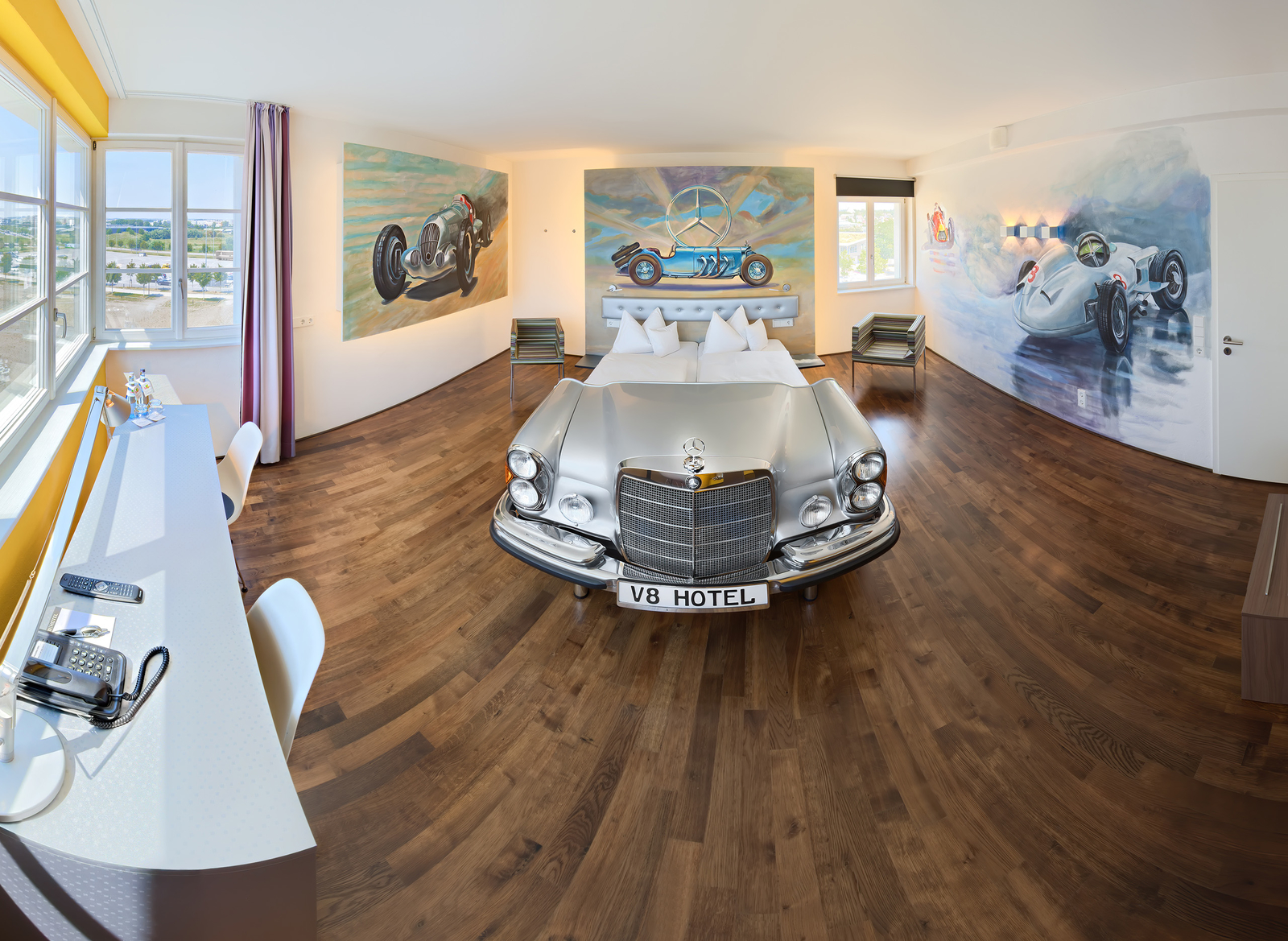 Das Bild zeigt ein Zimmer in einem Hotel, in dem ein silbernes Auto als Bett dient. An den Wänden hängen lebendige Wandmalereien von Oldtimern. Der Raum hat Holzböden und große Fenster, die Tageslicht hereinkommen lassen. Ein Schreibtisch und Stühle sind ebenfalls sichtbar.