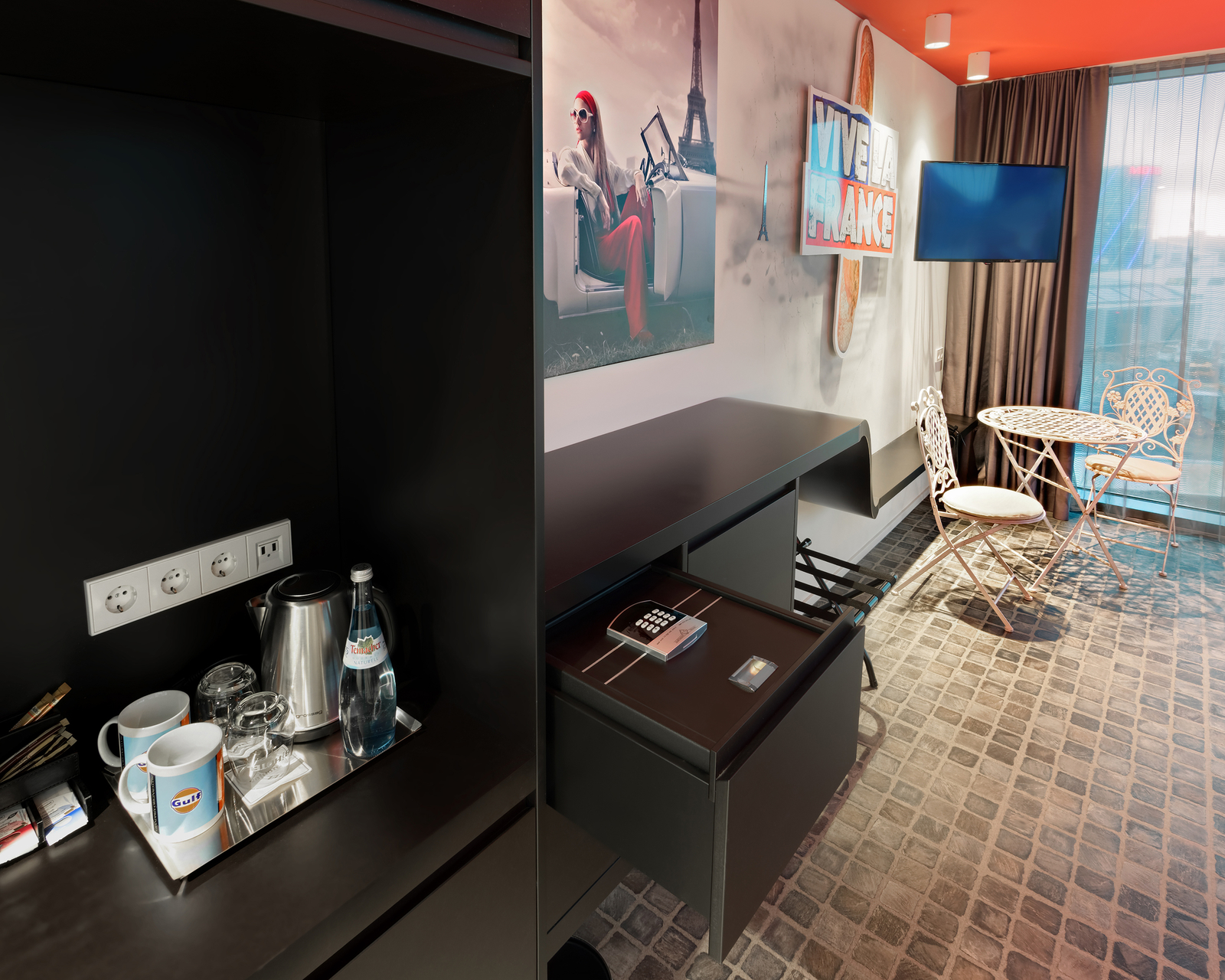 Das Bild zeigt eine stilvoll eingerichtete Hotelzimmer-Ecke. Zu sehen sind ein kleiner Tisch mit zwei Stühlen, ein Schreibtisch mit einem Sicherheitsschloss und eine Minibar mit Wasserflaschen und Tassen. An der Wand hängt ein Fernseher und ein dekoratives Wandbild mit einem Pariser Motiv.