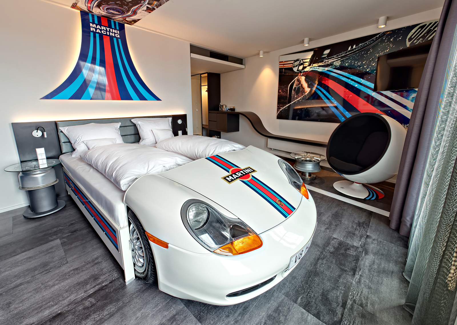 Weißes Autobett unter dem Martini Racing Logo gegenüber eines runden Sessels unter eines Fernsehers im Martini Racing-Themenzimmer im V8 Hotel