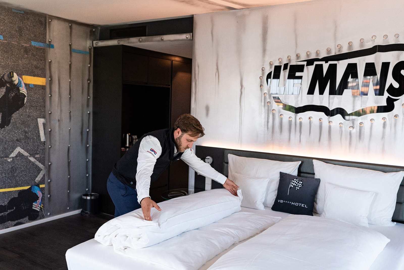 Ein Mitarbeiter macht das Bett in einem modern eingerichteten Hotelzimmer. Auf der Wand ist das Logo „LE MANS“ zu sehen. Die Bettwäsche ist weiß und ordentlich gefaltet, während der Mitarbeiter eine Decke zurechtlegt. Das Zimmer wirkt sauber und einladend.