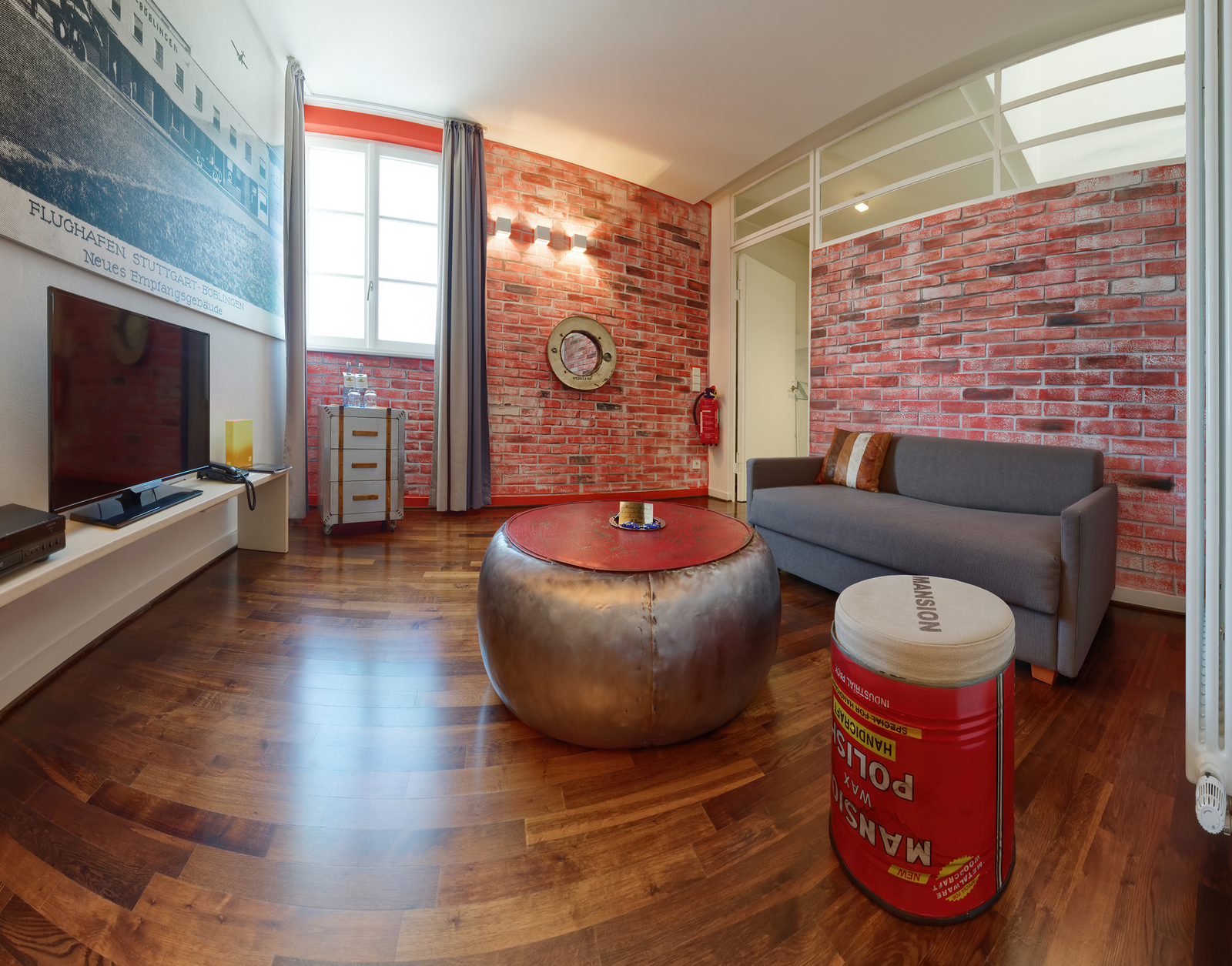 Der Raum ist modern eingerichtet mit einer grauen Couch und einem runden Tisch in der Mitte. Die Wände sind mit roten Ziegelsteinen dekoriert. Ein Fernseher steht auf einem Schrank und ein Fenster sorgt für Tageslicht. Ein kleines, rundes Tischchen ist ebenfalls vorhanden.