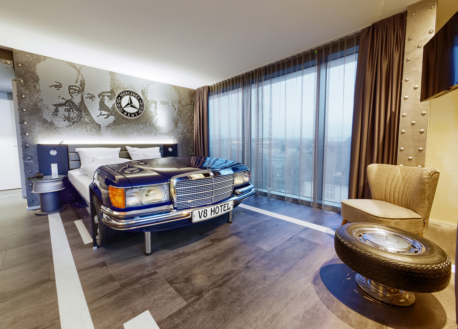 Blaues Mercedes Autobett neben einer Fensterfront gegenüber eines Sessels und Tischs aus einem Autoreifen im Mercedes Themenzimmer im V8 Hotel