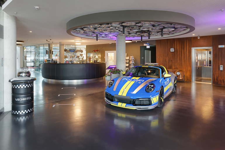 In der Empfangshalle eines Hotels steht ein blau-gelbes Sportauto mit Polizeidesign. Der Empfangstresen ist kurvenförmig und modern, im Hintergrund sind Glaswände und eine gemütliche Sitzecke. Die Beleuchtung sorgt für eine einladende Atmosphäre.