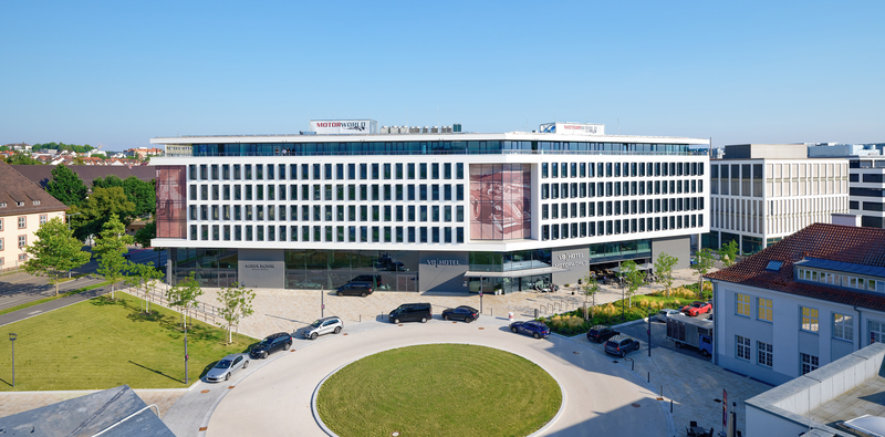 Das Bild zeigt ein modernes, mehrstöckiges Gebäude mit großen Fensterfronten. Im Erdgeschoss befindet sich der Eingang des „V8 Hotels“. Um das Gebäude herum sind grüne Flächen und mehrere parkende Autos zu sehen, während der Himmel klar und blau ist.