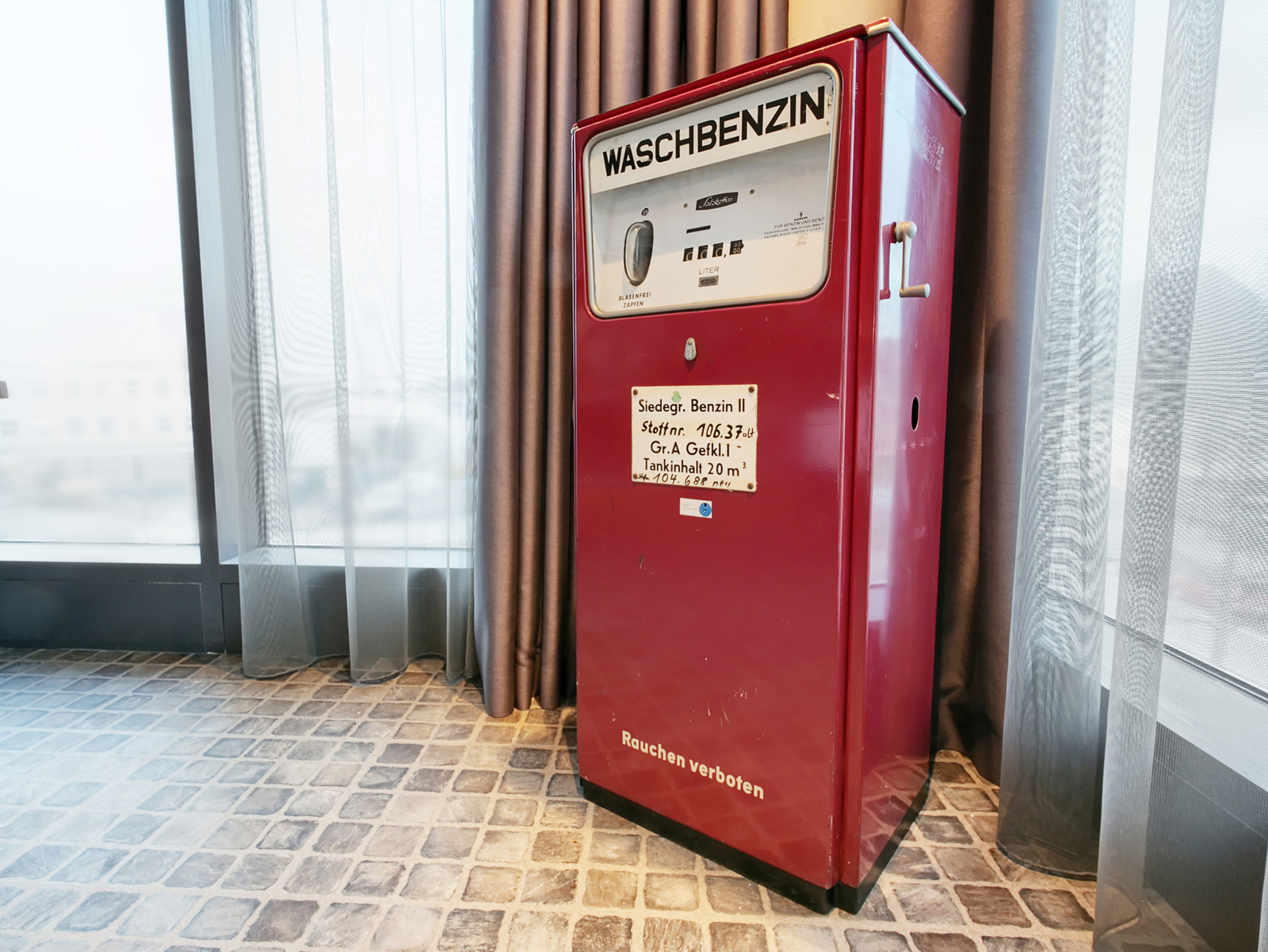 Ein roter Waschbenzin-Automat steht in einem Raum. Auf dem Automaten befindet sich eine informative Beschriftung über die Benzinsorte und technische Details. Neben dem Automaten ist ein Hinweis „Rauchen verboten“. Der Boden ist mit quadratischen Fliesen bedeckt.