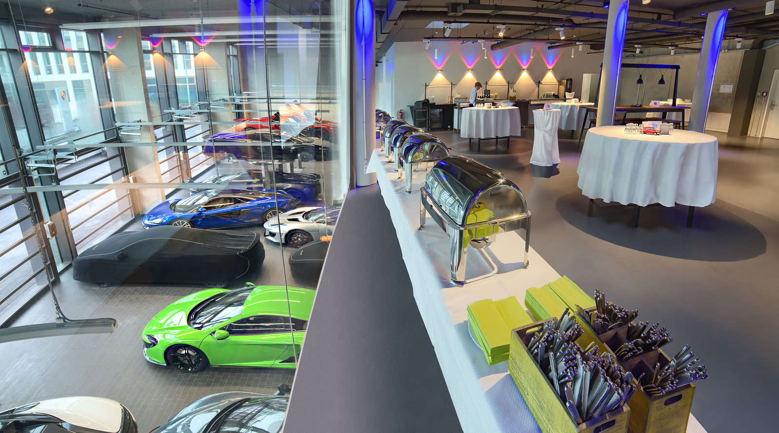 In einer modernen Eventlocation sind elegante Autos ausgestellt, während auf einem Tisch warme Speisen in großen, silbernen Chafing-Dishs präsentiert werden. Der Raum ist lichtdurchflutet und hat eine stilvolle Einrichtung mit runden Tischen und dekorativen Lichtern.