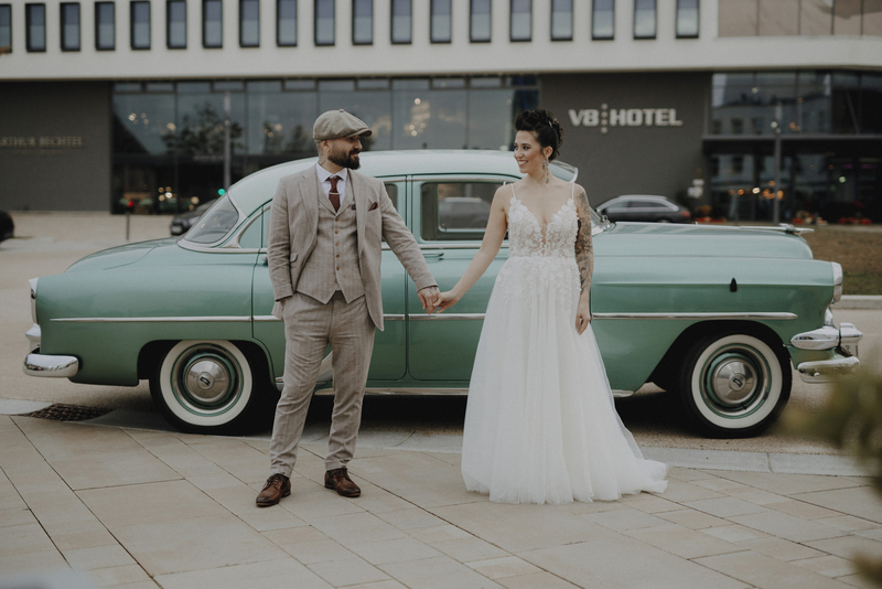 Eine Frau in Hochzeitskleid hält lächelnd die Hand ihres Mannes vor einem grünen Auto, im Hintergrund das V8 Hotel.